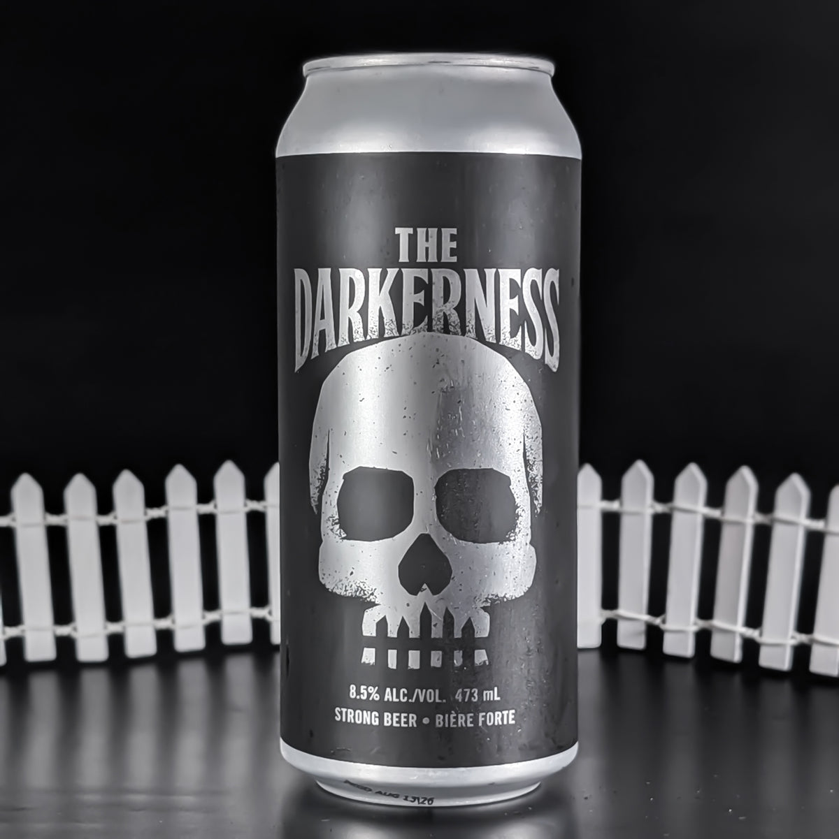 A 473 mL aluminum can of 'The Darkness' strong beer, featuring a large silver skull graphic on a dark gray label, set against a black background with a white picket fence.