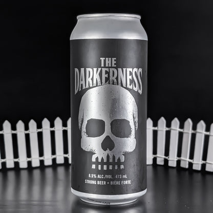 A 473 mL aluminum can of 'The Darkness' strong beer, featuring a large silver skull graphic on a dark gray label, set against a black background with a white picket fence.